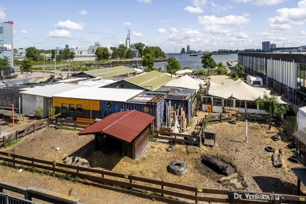 Urban farm "De VerbroederIJ" with animal enclosures and repurposed buildings along the Johan van Hasseltweg in Amsterdam, overlooking the IJ river.